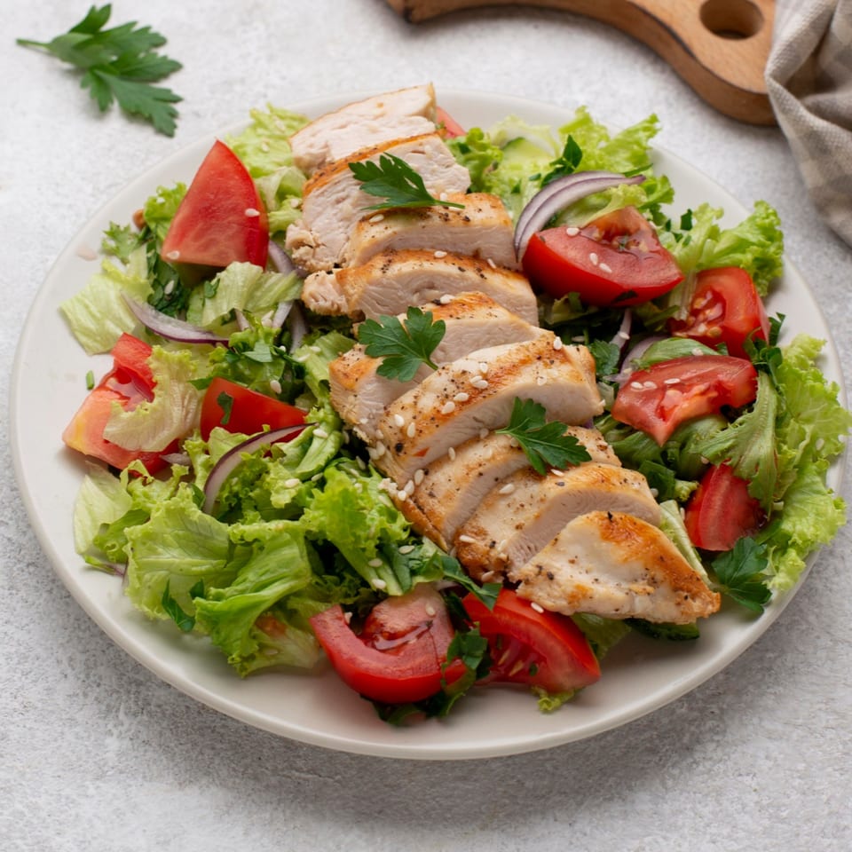 A fresh salad with sliced grilled chicken, lettuce, tomatoes, red onion, and sesame seeds.