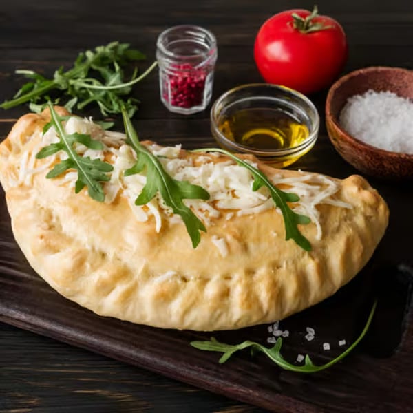 A golden-brown calzone topped with melted cheese and fresh arugula, alongside olive oil, salt, and a tomato.