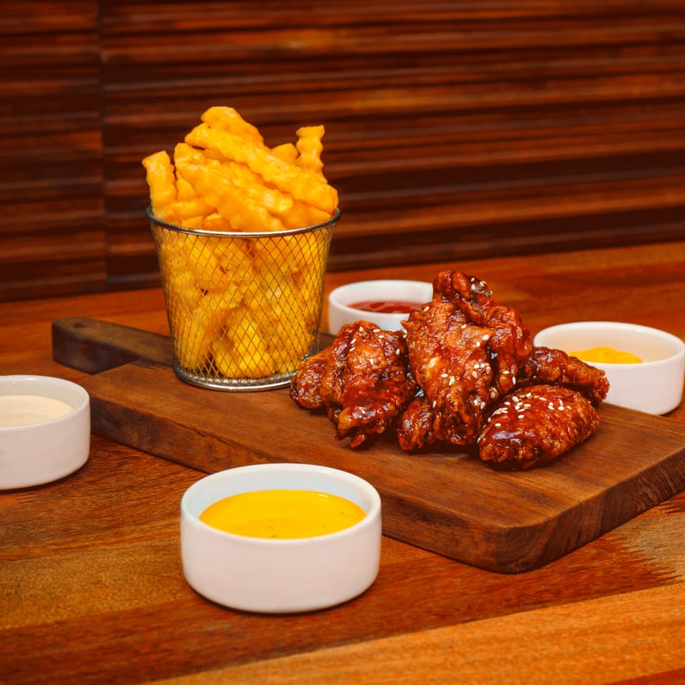 Crispy french fries in a metal basket, BBQ chicken wings, and dipping sauces on a wooden board.